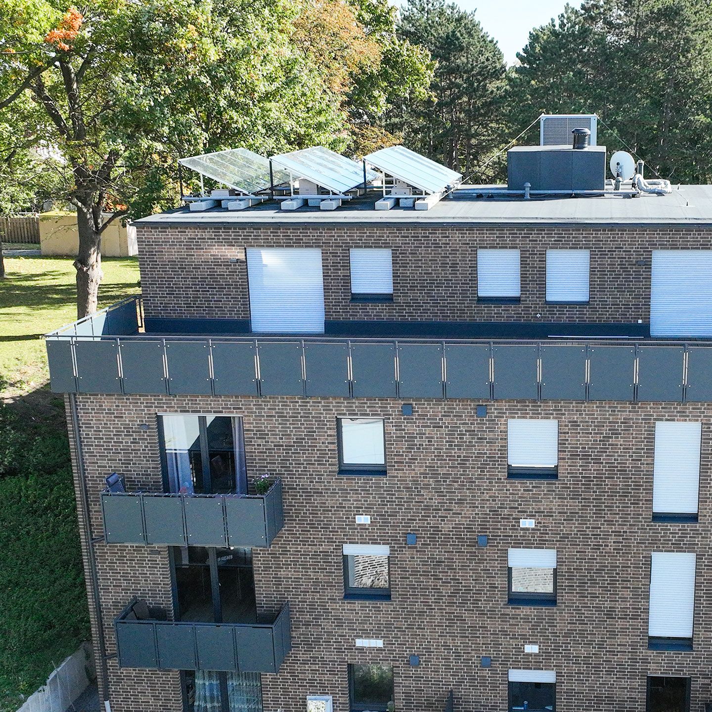 Ein modernes Wohngebäude mit brauner Ziegelfassade, Solarpaneelen auf dem Dach und umgeben von Bäumen.