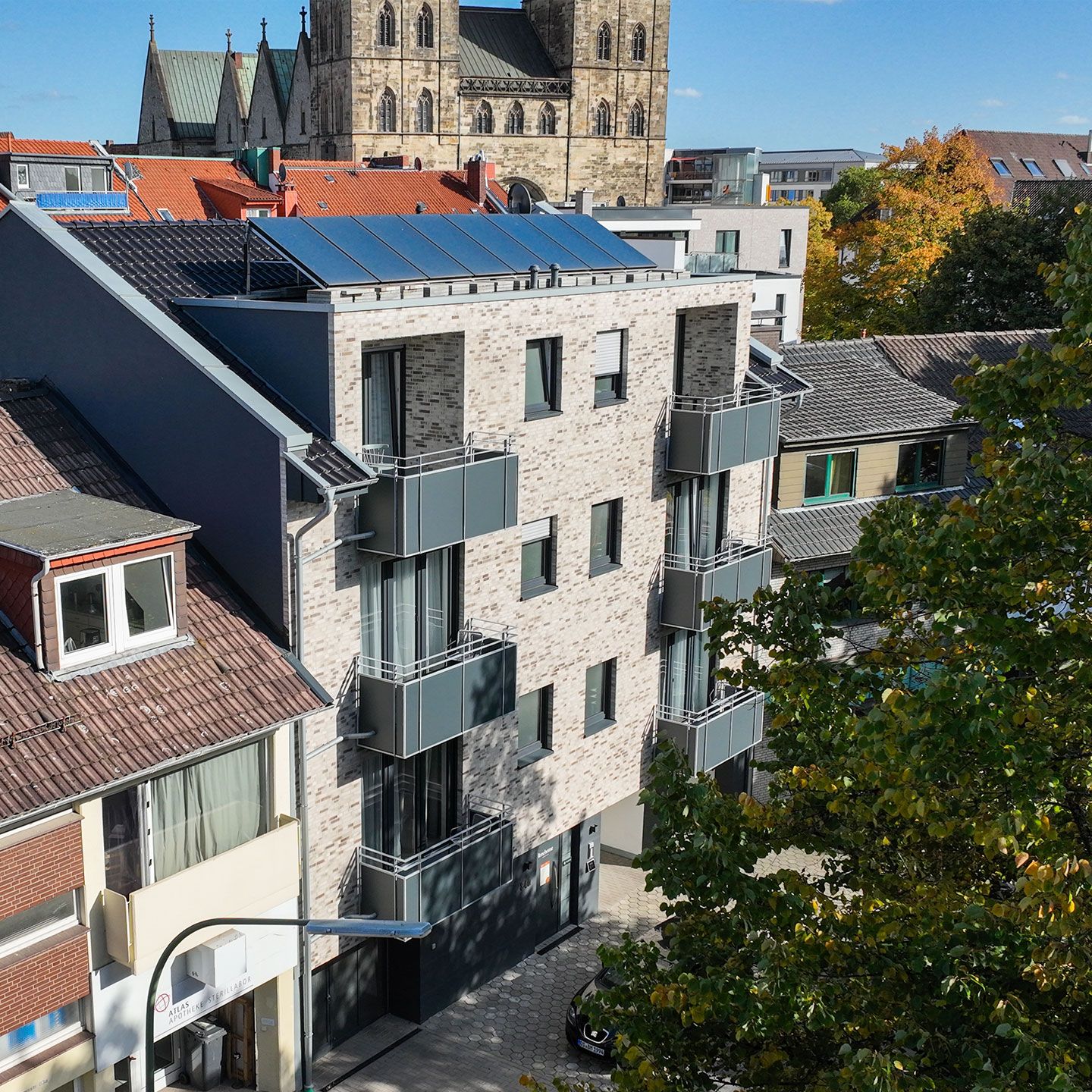 Moderne Gebäude mit Solarpanelen vor einer historischen Kirche, umgeben von grünen Bäumen.
