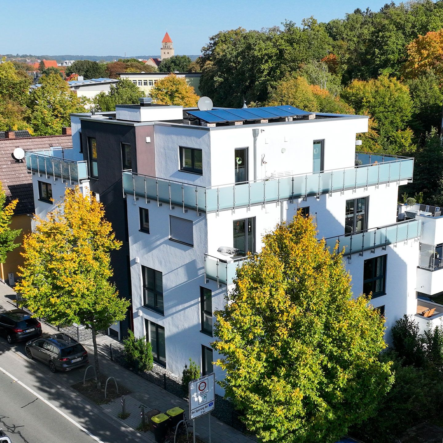 Modernes Wohngebäude mit Solarpanels, Glasbalkon und herbstlichen Bäumen in urbaner Umgebung.