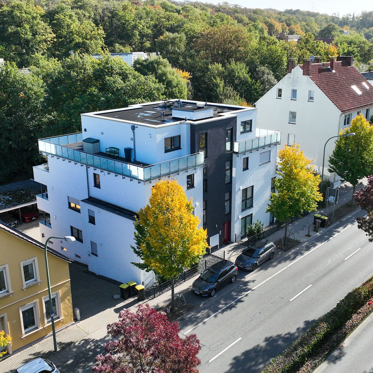 Modernes, mehrstöckiges Gebäude mit weißer Fassade, umgeben von herbstlichen Bäumen und parkenden Autos.