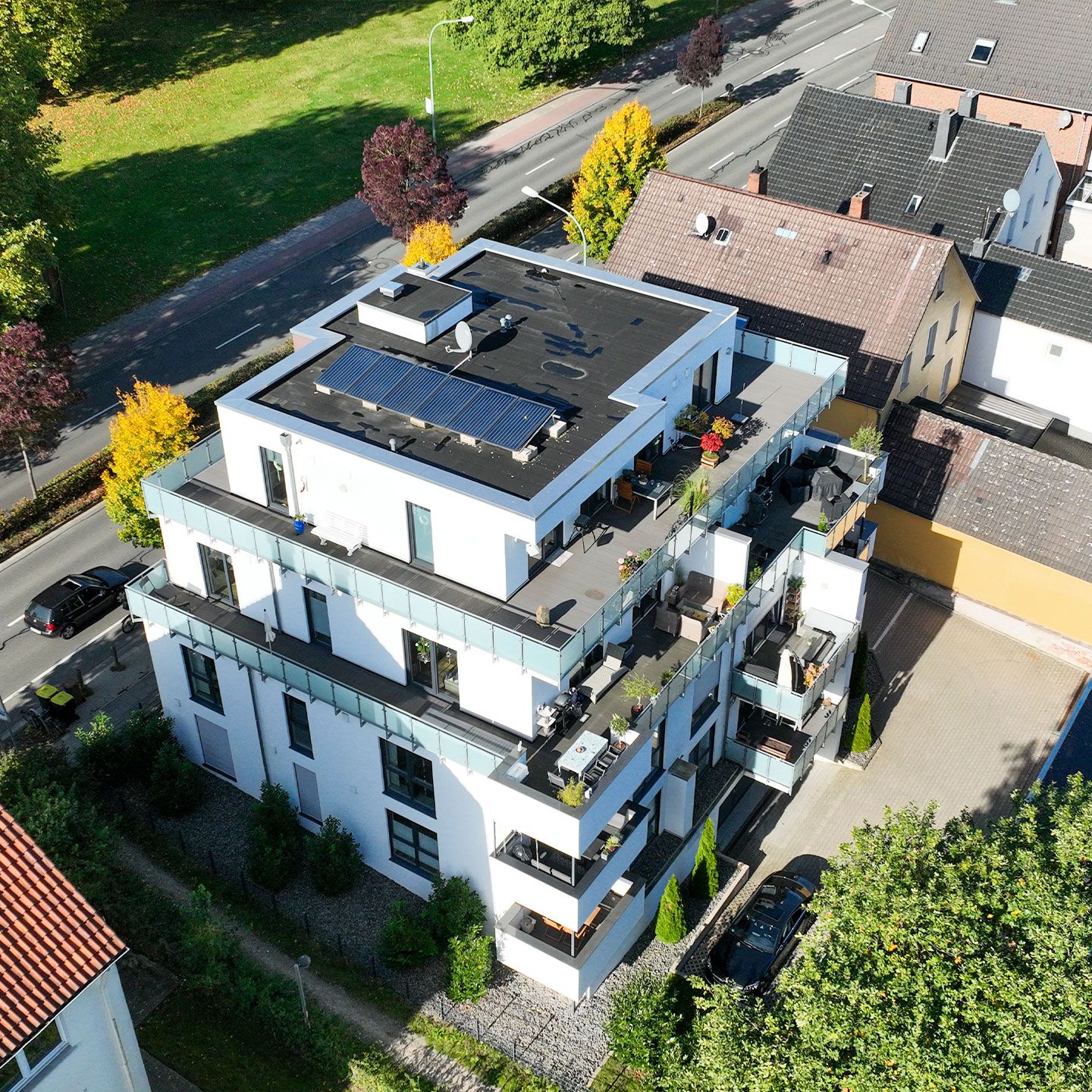 Moderne Wohnanlage mit Solarpaneelen auf dem Dach, umgeben von Bäumen und Autos auf der Straße.