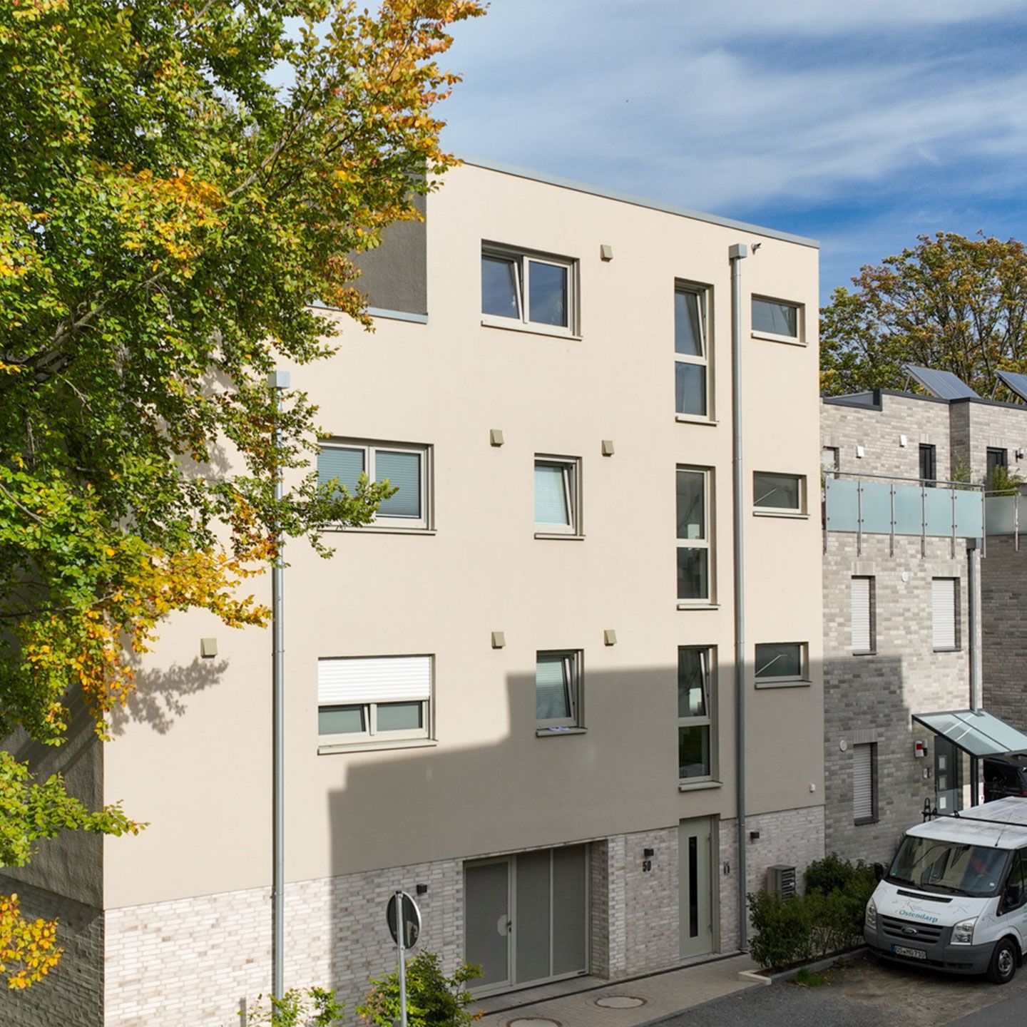 Modernes Wohngebäude im Herbst, mit hellen Fassaden, blauem Himmel und einem Baum mit buntem Laub.