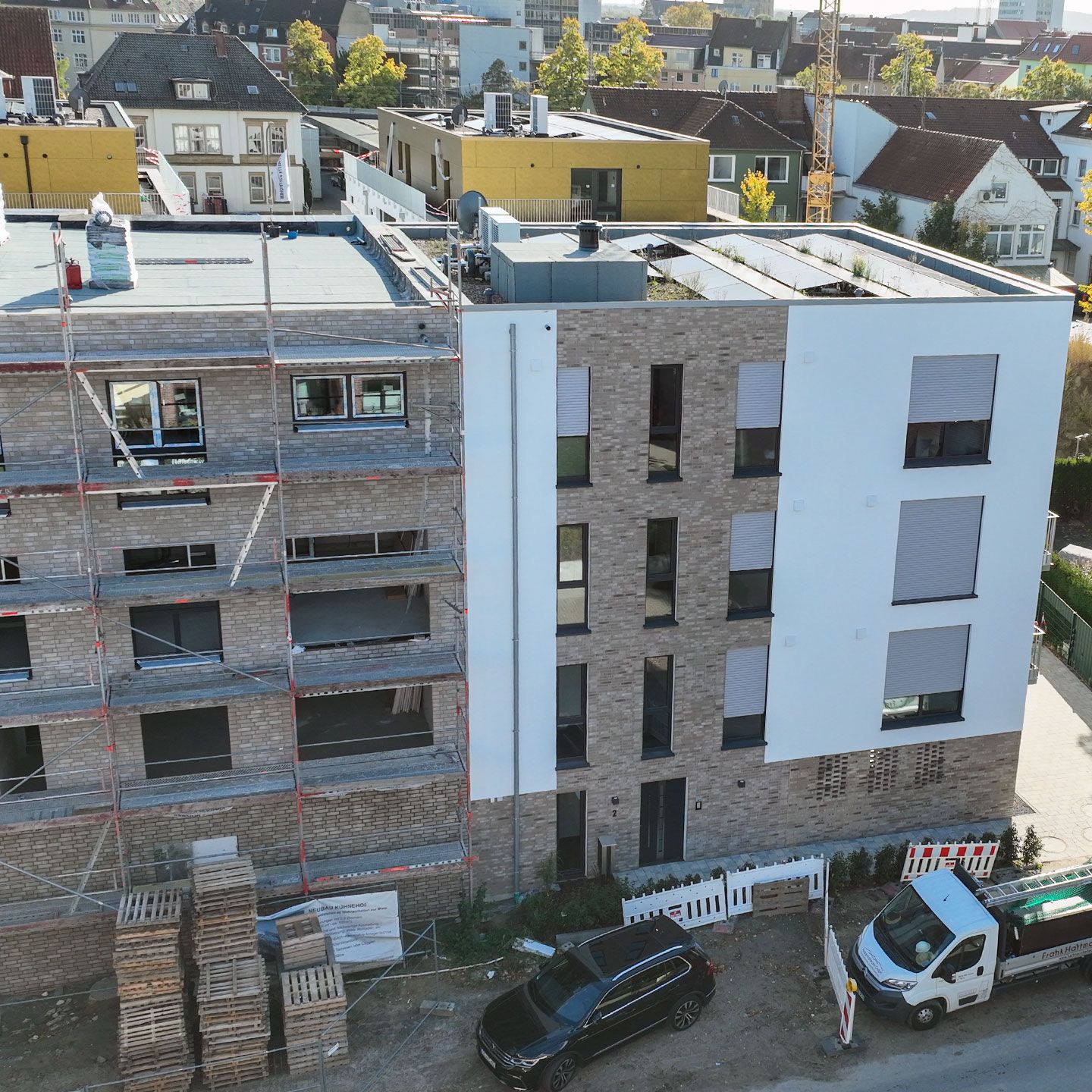 Städtische Baustelle mit Gerüst, Baumaterialien und geparkten Fahrzeugen neben einem fertigen Gebäude.