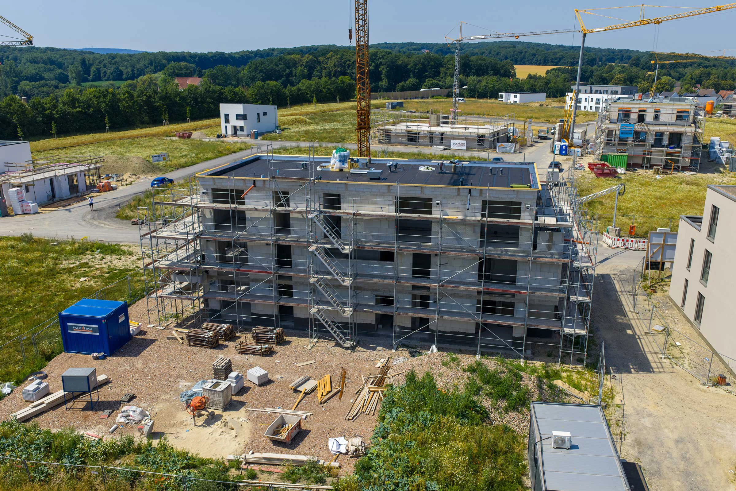 Eine ruhige Baustelle mit einem mehrstöckigen Rohbau, umgeben von Gerüsten und Baukränen in ländlicher Umgebung.