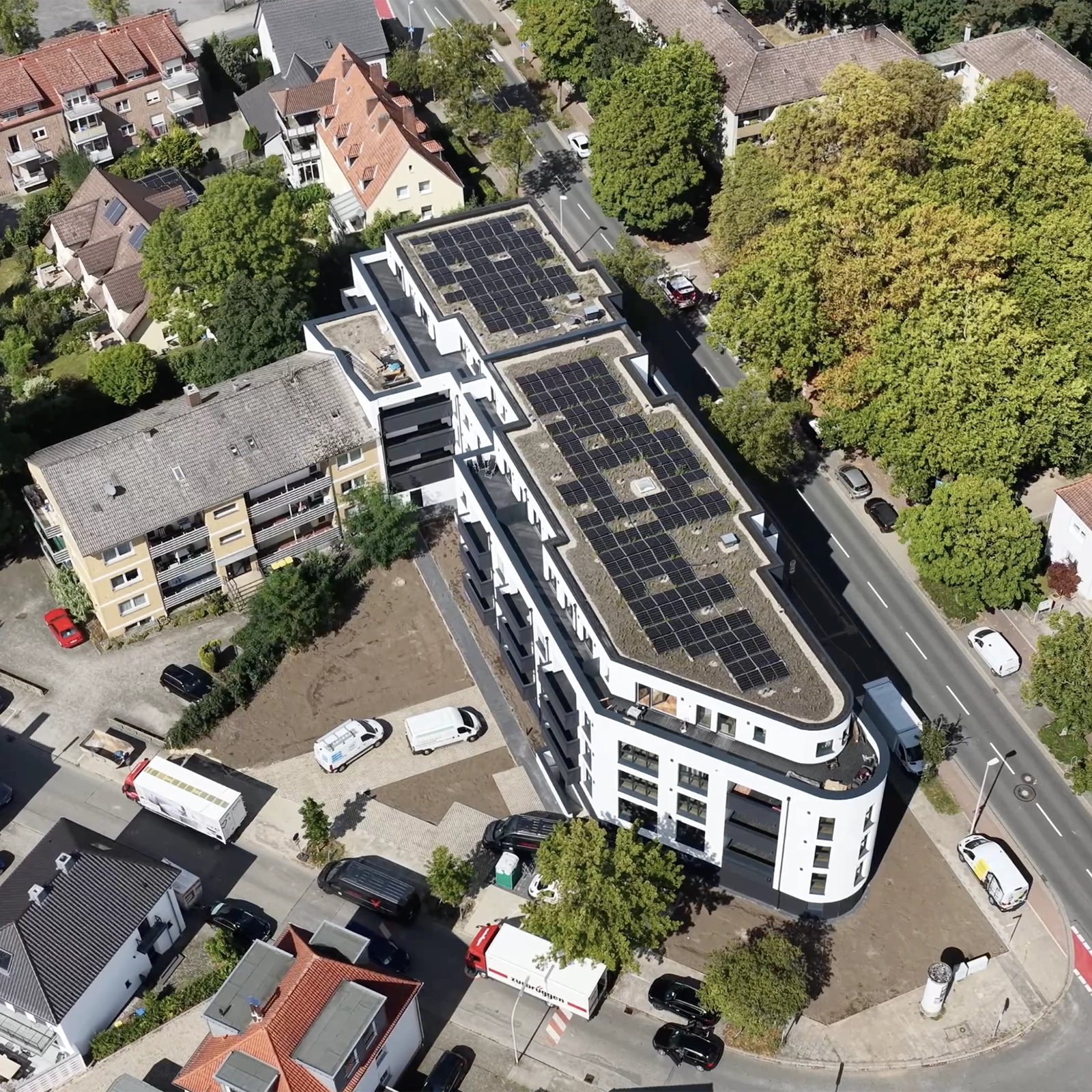 Luftaufnahme eines modernen, geschwungenen Gebäudes mit Solarpaneelen, umgeben von Wohnhäusern und Bäumen.
