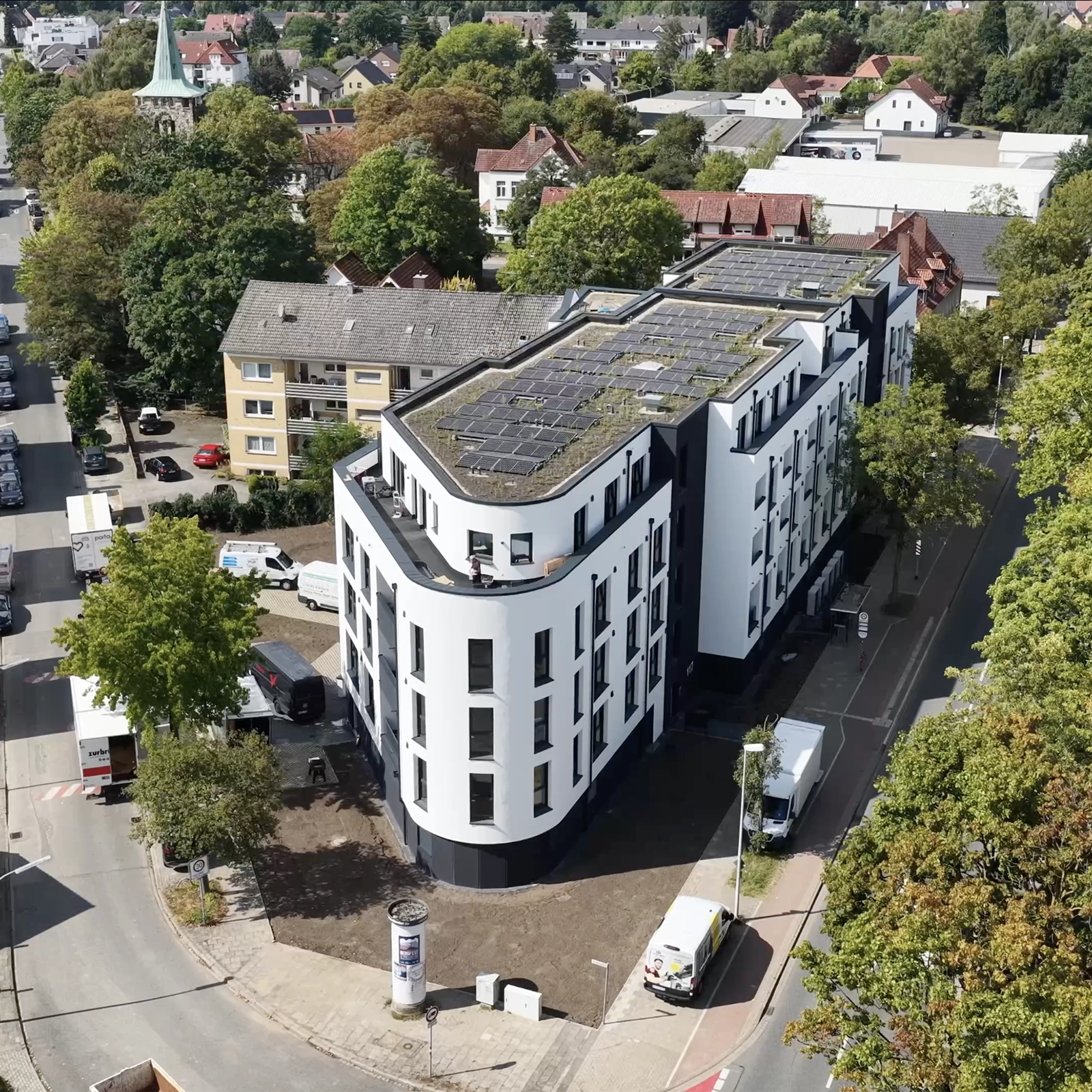 Vogelperspektive auf modernes Gebäude mit Solarpaneelen, umgeben von geparkten Autos und älteren Häusern.