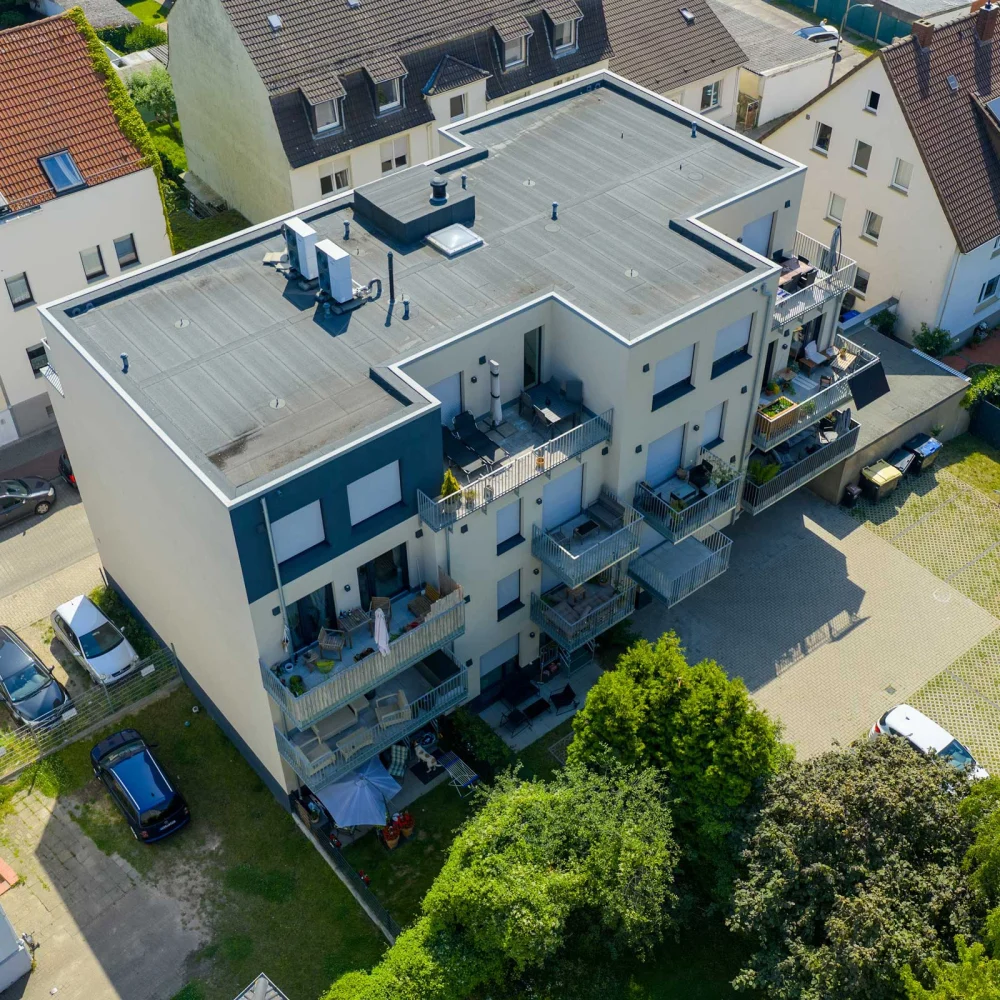 Vogelperspektive auf modernes Mehrfamilienhaus mit Balkonen, umgeben von Gärten und geparkten Autos.
