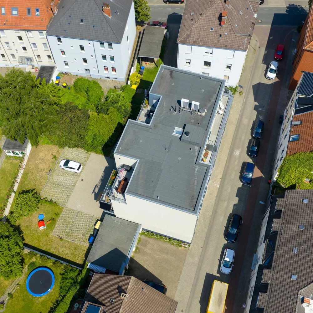 Luftaufnahme einer ruhigen Wohngegend mit bunten Dächern, grünem Garten und einem weißen Auto.