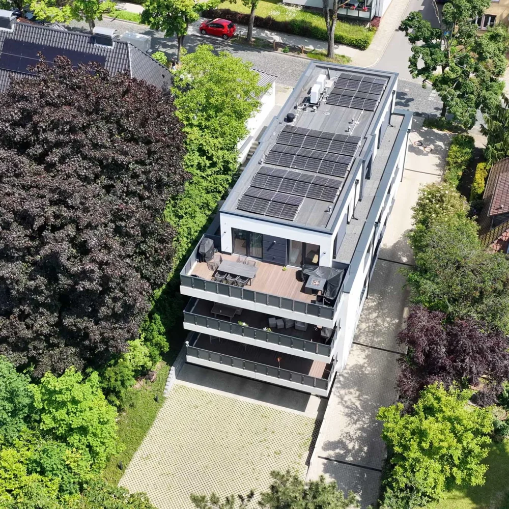 Luftaufnahme eines modernen Wohngebäudes mit Solarpaneelen, umgeben von grünen Gärten und Bäumen.