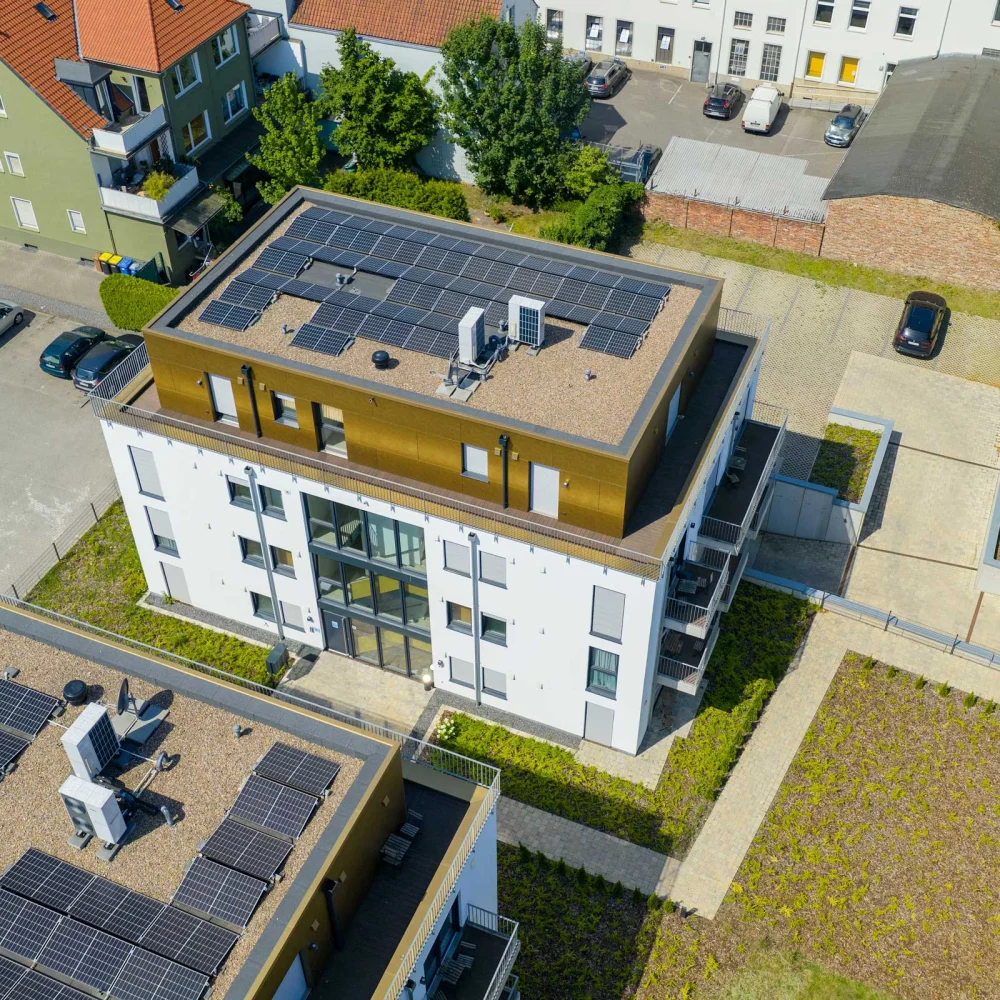 Luftaufnahme eines modernen Wohngebäudes mit Solarpaneelen auf dem Flachdach, umgeben von Grünflächen.