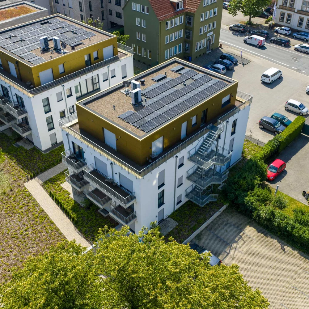 Moderne Wohngebäude mit Solarpaneelen, umgeben von Bäumen und geparkten Autos in einer ruhigen Straße.
